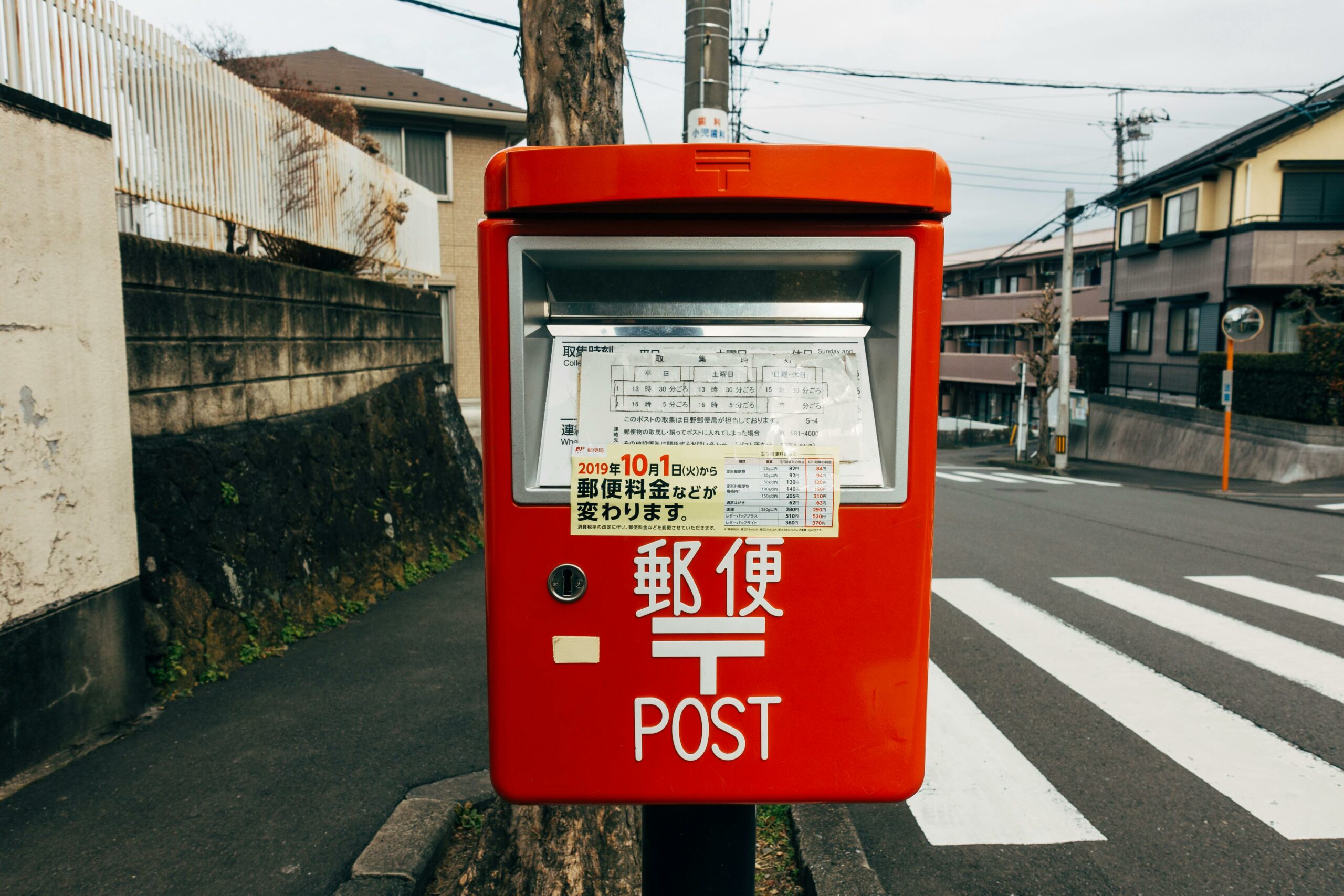 Bright red mail postbox in a Japanese town, with a crosswalk and urban architecture in the background.
